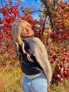 a beautiful blonde woman standing in front of some trees with red and yellow leaves on it