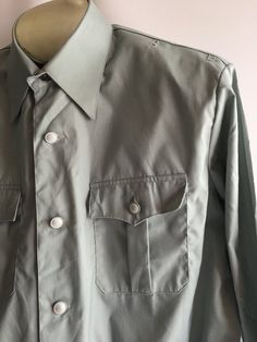 Awesome vintage men's uniform work shirt jacket. The military inspired cut and color encourage me to believe this was a aviation uniform of some sort. Somewhere between a shirt and a jacket, these piece has some great details. The material is most likely a permanent press style poly/cotton blend, though no content label is present. The shirt has a button front styling much like a classic men's dress shirt but with a more boxy cut and elastic waist. It has small loops and holes near top shoulder Military Style Button-up Work Shirt, Military Style Button-up Shirt For Work, Military Style Workwear Button-up Shirt, Military Style Collared Shirt For Workwear, Military Style Shirt For Work With Button Closure, Military Style Shirt With Buttons For Work, Military Style Workwear Shirt With Buttons, Military Long Sleeve Work Shirt, Military Style Workwear Tops With Snap Buttons