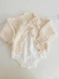 Soft and cozy, this stunning knit cardigan is the perfect addition to your little ones closet. Tiny Pom Pom appliqués all over. Textured pattern with front buttons. Playful Knit Long Sleeve Outerwear, Playful Knit Cardigan For Fall, Playful Cotton Knitted Cardigan, Playful Knitted Cotton Cardigan, Playful Knit Cardigan For Winter, Cute Long Sleeve Knit Cardigan, Cozy Long Sleeve Sweater For Playtime, Cream Cotton Cardigan With Buttons, Cute Cream Cotton Cardigan