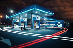 an empty gas station at night with the lights streaking across the road and cars passing by