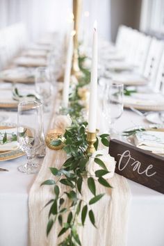 the table is set with white linens and greenery on it, along with candles