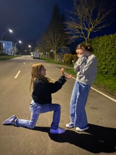 two people standing in the middle of an empty road at night, one kneeling down to touch the other's hand