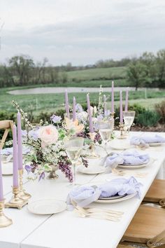 a long table with purple candles and flowers on it is set for an outdoor dinner