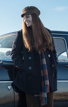 a woman standing in front of a black car wearing a hat and scarf with her hands on her hips