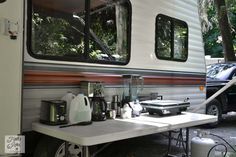 a camper parked in the woods with its door open and coffee pot on the table next to it