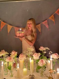 a woman standing in front of a table filled with flowers and candles holding a cake