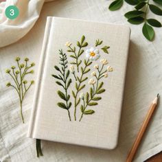 an embroidered book with flowers on it next to a pencil and some green leafy plants