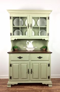 a green china cabinet with glass doors and plates on the top, in front of a white wall