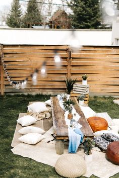 an outdoor picnic is set up on the grass in front of a wooden fence with pillows and blankets