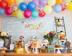 a table topped with lots of balloons next to a wall filled with cake and desserts