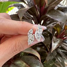 a woman's hand holding two diamond earrings in front of a potted plant