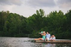 a family sitting on a dock in the middle of a lake with trees behind them