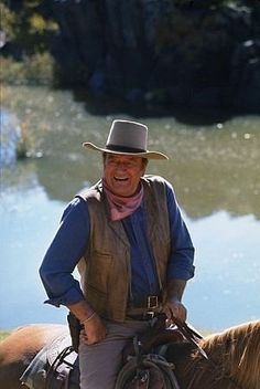 an older man riding on the back of a brown horse next to a lake and forest