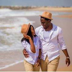 a man and woman are walking on the beach with their hats over their heads while looking at each other