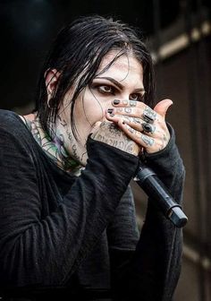 a man with black hair and piercings holding his hands to his mouth while standing in front of a microphone