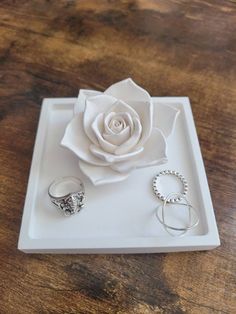 a white rose sitting on top of a wooden table next to two silver rings and a ring holder