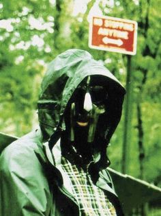 a man wearing a mask and raincoat standing in front of a sign with trees