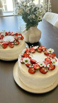 two cakes decorated with strawberries and daisies sit on a table in front of a vase