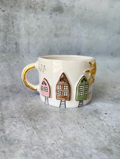 a coffee cup with houses painted on the side and gold rimmed handles, sitting on a gray surface