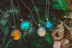 three necklaces with different designs on them sitting next to some green leafy plants