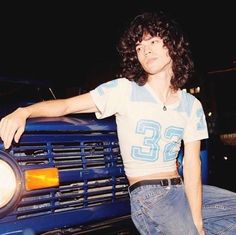 a woman leaning on the front grill of a blue truck with her hand on it's hip