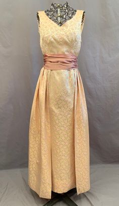 "Vintage 1960s formal in lovely pink and gold on cream brocade.  it is really two dresses in one as it is a long fitted sheath with a separate full overskirt plus cumberbund / sash.  Dress is sleeveless with a scoop neck that is lower in back, side zipper and long back kick pleat.  Skirt is set to a narrow waist band that fastens to dress with hooks and eyes.  It gathers across the back, wrapping part way over the front ending in soft pleats. The rose satin sash has no fastenings -- fold it over Elegant Brocade Dress For Occasion Wear, Gold Dress With Pleated Bodice For Wedding, Gold Wedding Dress With Pleated Bodice, Fitted Brocade Gown For Formal Occasions, Formal Brocade Gown With Fitted Bodice, Vintage A-line Wedding Evening Dress, Brocade Dresses With Fitted Bodice For Evening, Gold Fitted Bodice Dress For Mother Of The Bride, Elegant Beige Vintage Dress For Evening