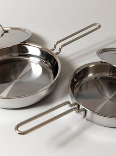 three stainless steel pots and pans on a white surface with one holding an empty frying pan