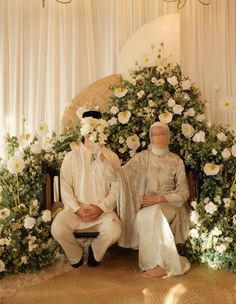 two men sitting next to each other in front of a flower covered arch with white flowers