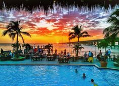 people are sitting at tables next to the pool as the sun sets over the ocean