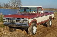 a red and white truck driving down a dirt road next to a body of water