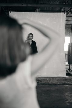 a woman standing in front of a white wall with her hands behind her head looking into the mirror