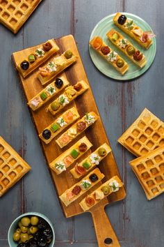 waffles and olives are arranged on a wooden cutting board next to plates with different types of appetizers