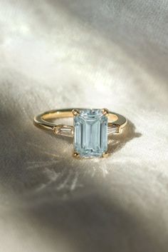 a ring with an aquamarinee stone and two white diamonds on the side, sitting on a silver cloth