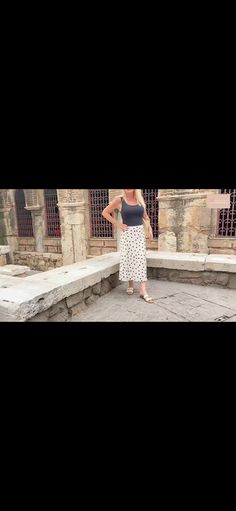 a woman standing in front of a stone wall with her hands on her hips, wearing a black tank top and white polka dot skirt