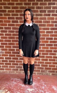 a woman in black dress and boots standing next to a brick wall with her tongue out