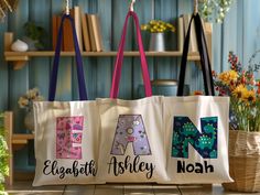 three personalized totes hanging from hooks in front of bookshelves and flowers