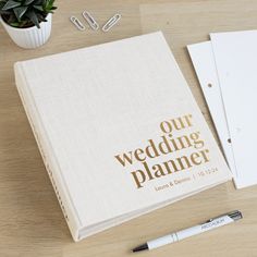 a wedding planner next to a pen, notebook and flower pot on a wooden table