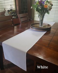 a white table runner on top of a wooden dining room table with flowers in a vase