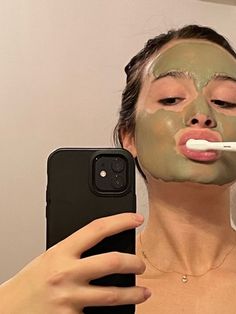 a woman brushing her teeth with a green mask on while looking at her cell phone