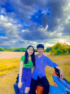 a man and woman sitting on top of a blue motorcycle in a grassy field under a cloudy sky