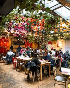 many people are sitting at tables in a restaurant with hanging plants and flowers on the ceiling