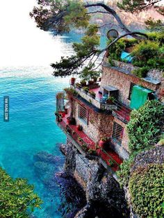 an old house on the edge of a cliff by the ocean with plants growing out of it