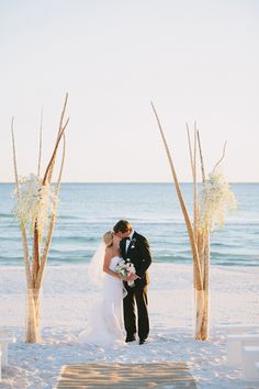 a bride and groom kissing on the beach in front of an arch made out of branches