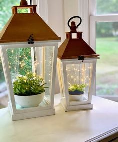 two white lanterns with plants in them sitting on a window sill