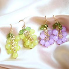 three different colored grapes are hanging from gold earwires on a white satin surface