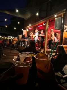 two people holding drinks in their hands while sitting on a bench outside at night time