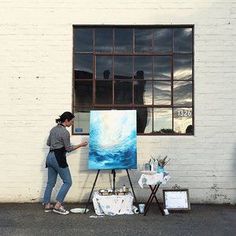 a woman standing next to an easel with a painting on it in front of a window