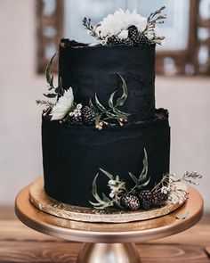 a three tiered black cake with white flowers and greenery on top sits on a gold plate