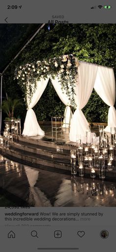 an outdoor wedding setup with white drapes and flowers on the stage, surrounded by candles