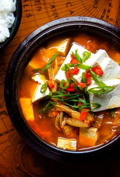 a bowl filled with tofu and vegetables on top of a wooden table next to rice